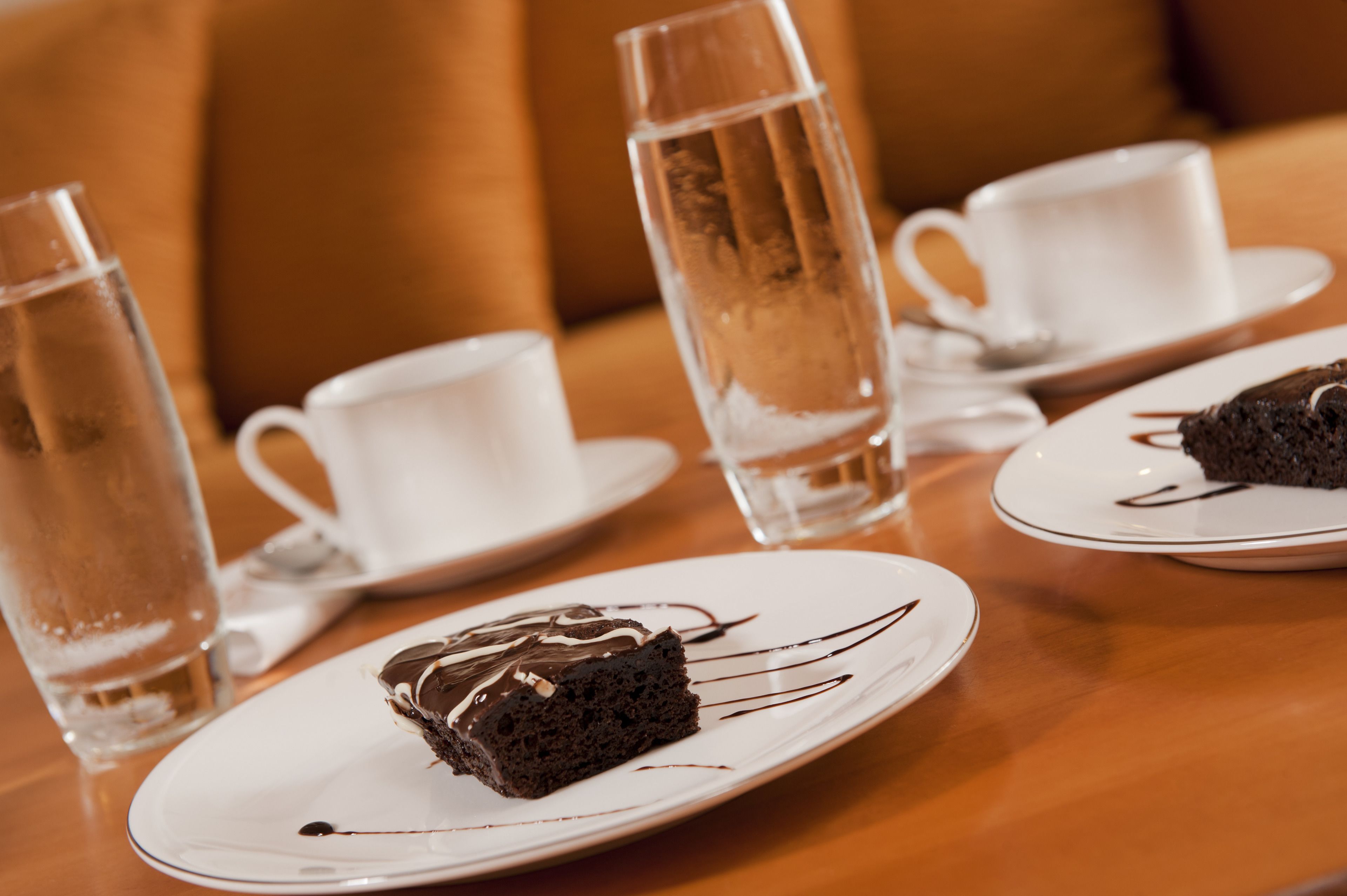 A table with a slice of chocolate cake on a white plate, a tall glass of water, and a white cup on saucer.