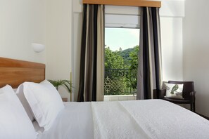 A hotel room with a bed, a chair, a window with curtains, and a view of greenery outside.