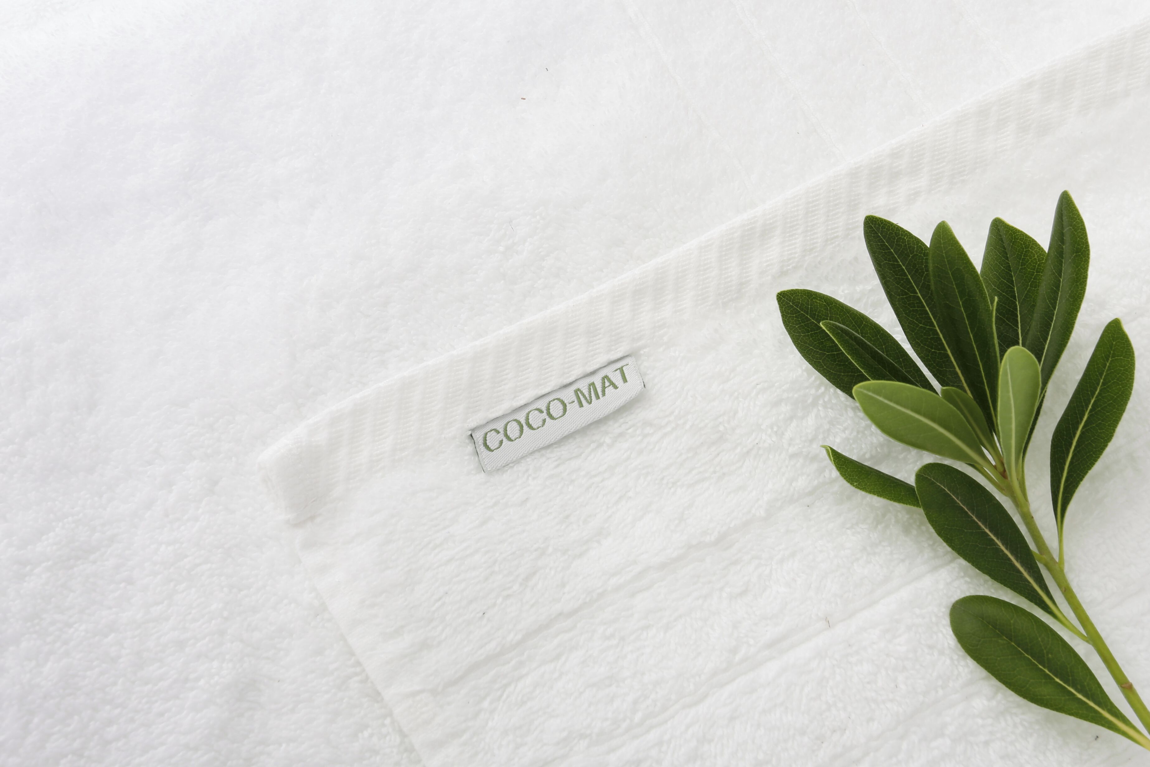 A white textured surface with a label reading "COCO-MAT" and a branch of green leaves.