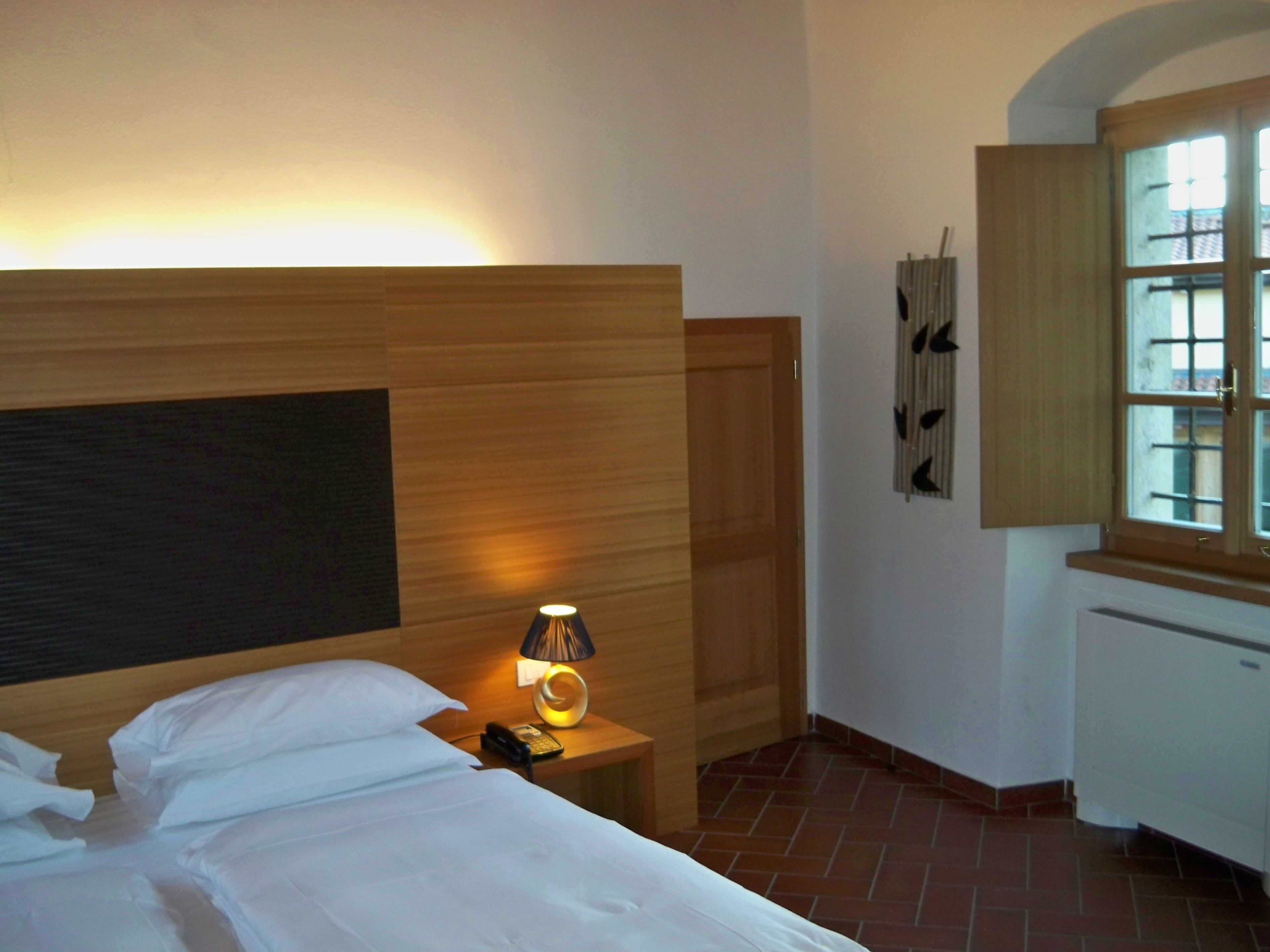 Ein Hotelzimmer mit einem Bett, einem Nachttisch mit Lampe, einem Holz-Kopfteil, einem Fenster mit Vorhängen und einer kleinen Küchenzeile.