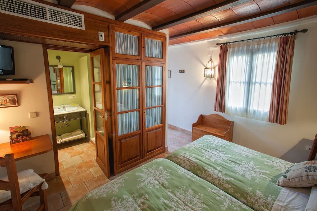 Un dormitorio con dos camas, un armario de madera, un baño con lavamanos y un televisor sobre una estantería.
