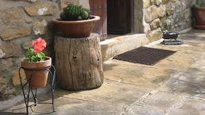 Un muro de piedra con una puerta de madera, una planta en maceta, un banco de metal y una rejilla metálica sobre un suelo de piedra.