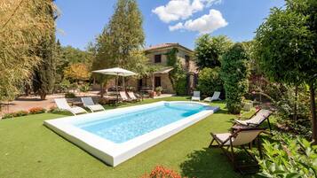 Una piscina con sillones de descanso, una casa de piedra y abundante vegetación.
