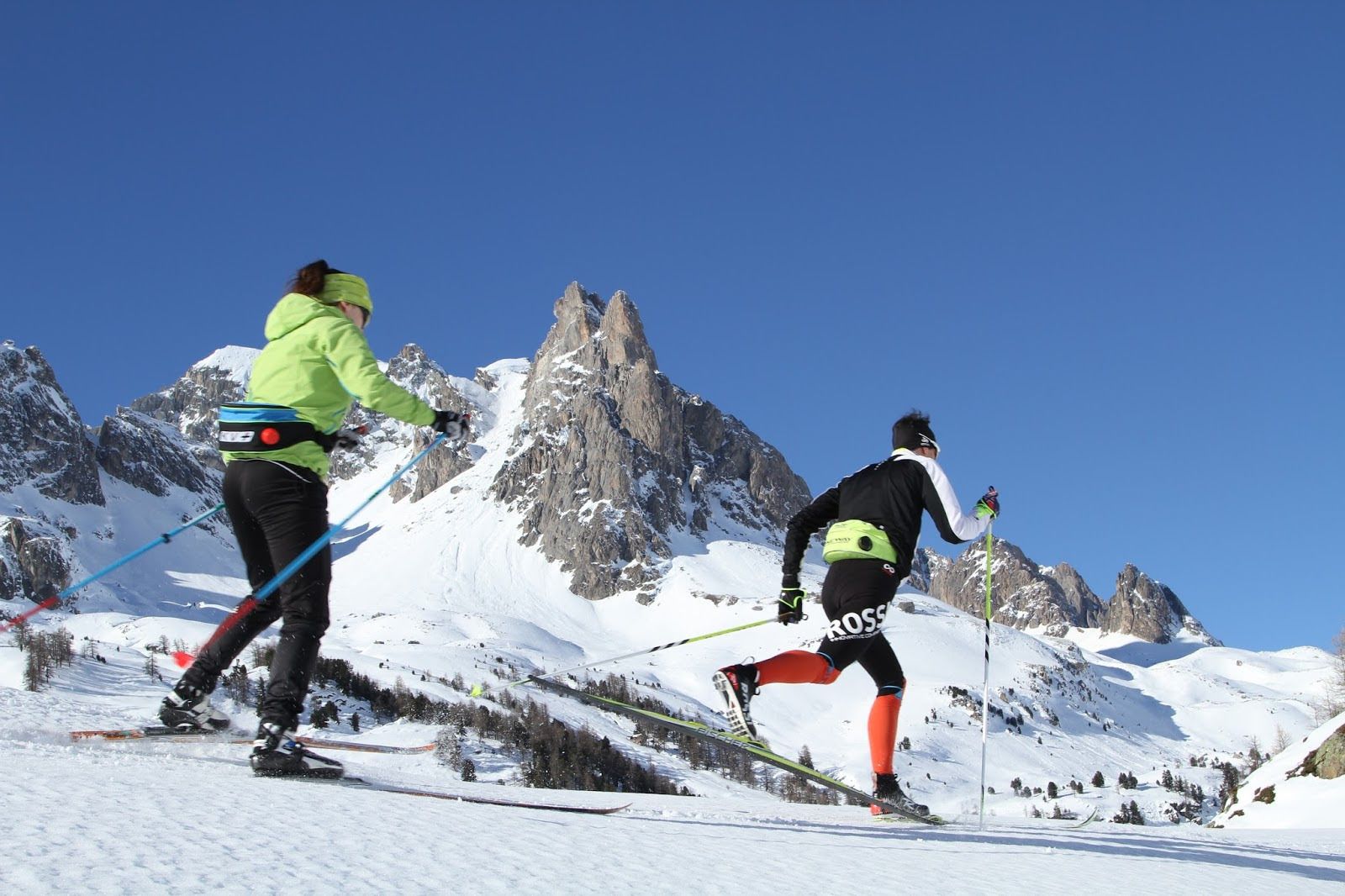 Two skiers on a snowy mountain slope with rocky peaks in the background.