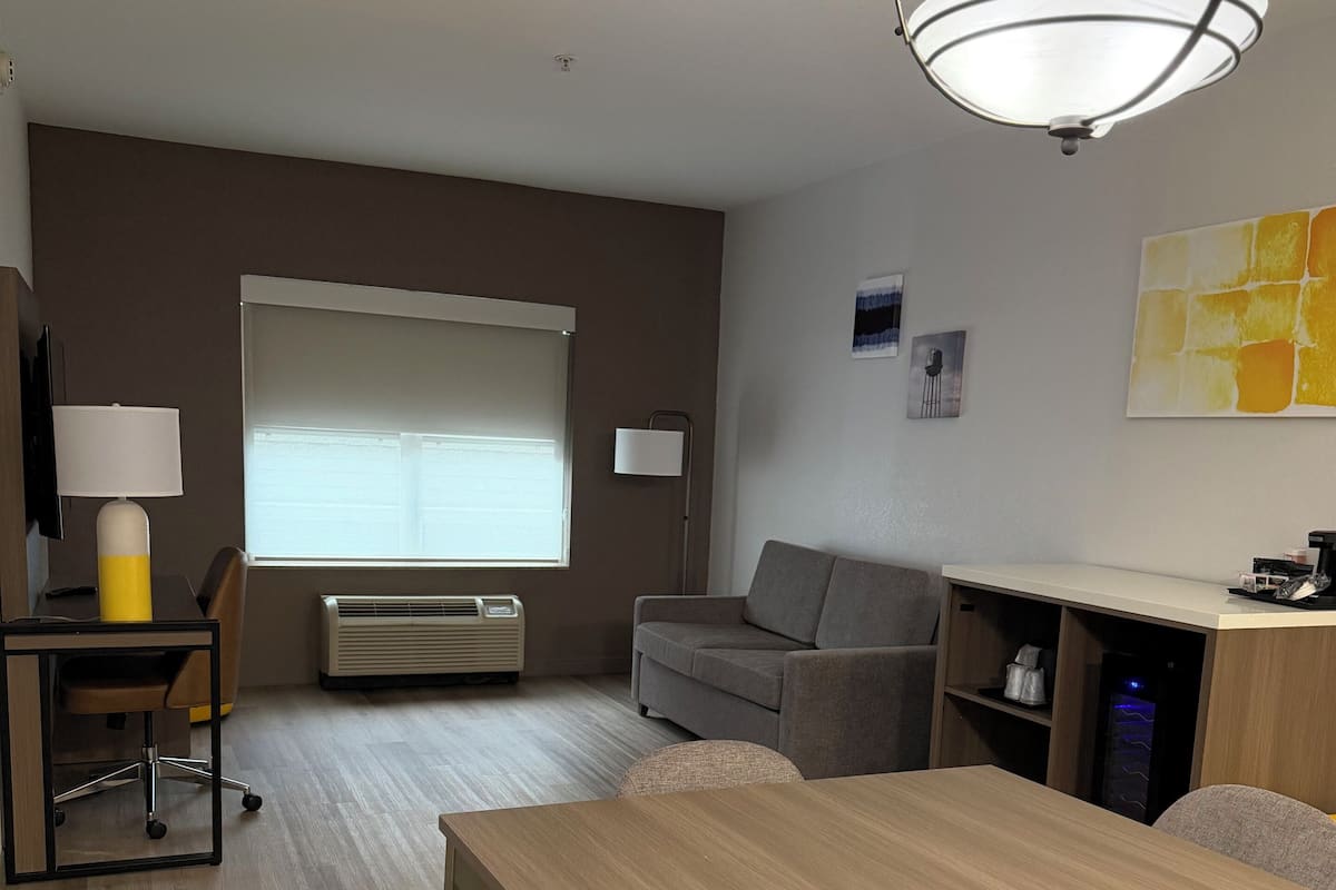 Ein modernes Hotelzimmer mit Esstisch aus Holz, grauem Sofa und großem Fenster mit Jalousien.