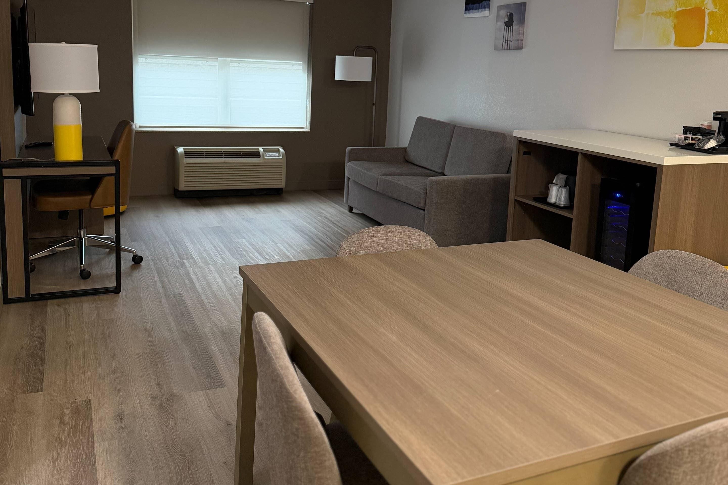 A modern hotel room with a wooden table, a sofa, a desk with a lamp, an air conditioning unit, and a bookshelf.