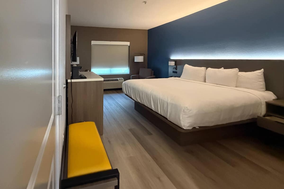 Ein Hotelzimmer mit Bett, Schreibtisch, Stuhl, Sitzbank, Fernseher und Fenster mit Jalousien.