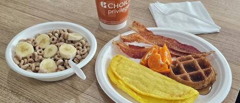 A breakfast plate with waffles, bacon, orange slices, and a bowl of cereal with banana slices.