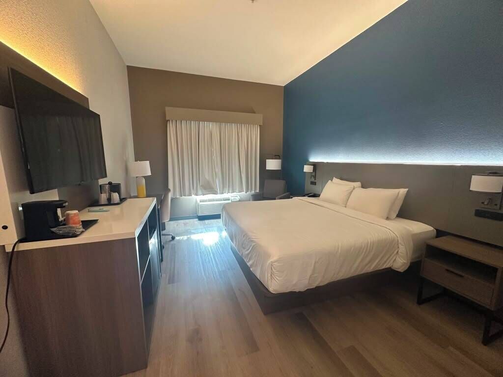 Hotellihuone, jossa on sänky, yöpöytä, lamppu, televisio ja työpöytä tuolilla.