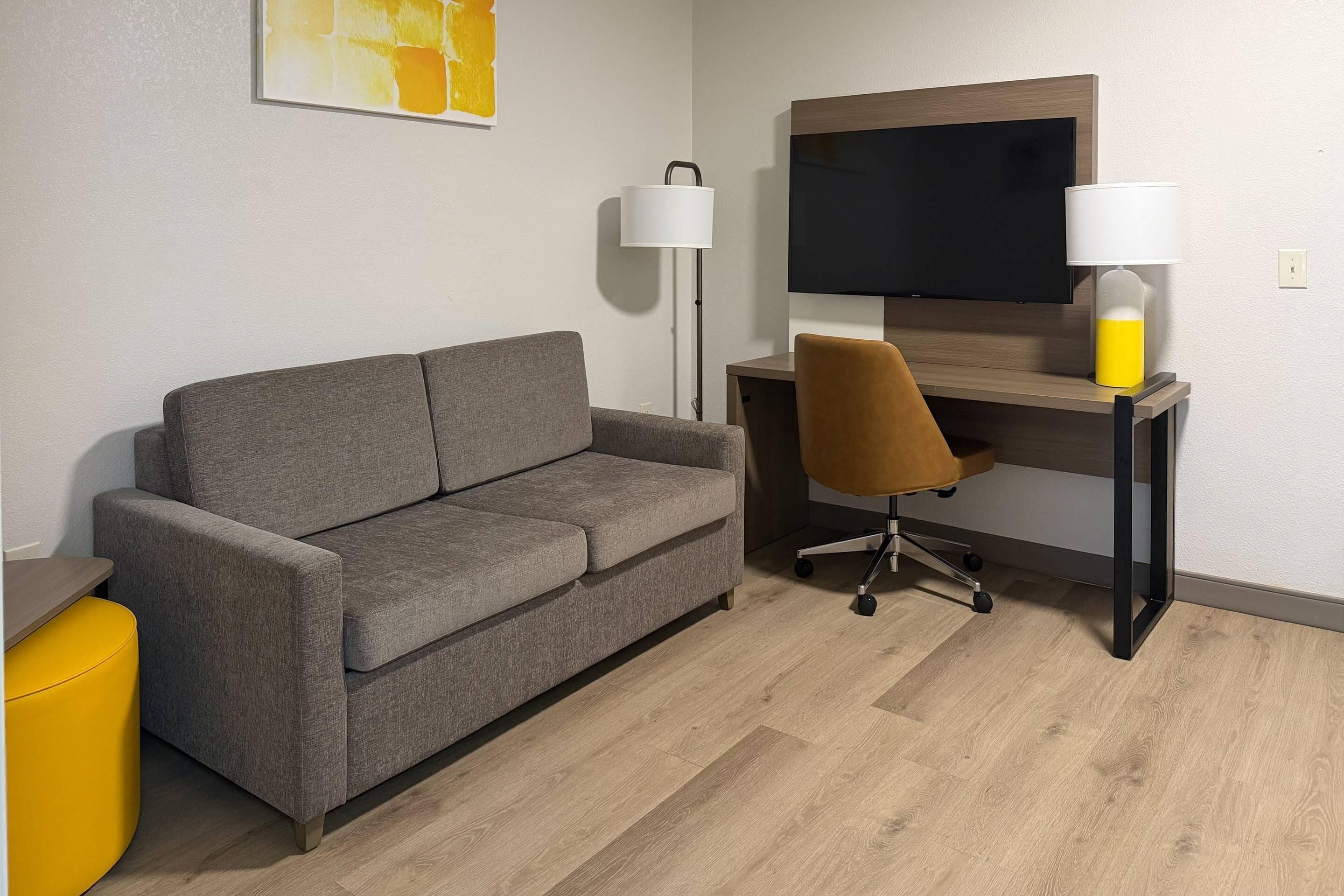 A modern room with a grey sofa, a wooden desk, a brown office chair, a yellow ottoman, and a flat-screen TV.