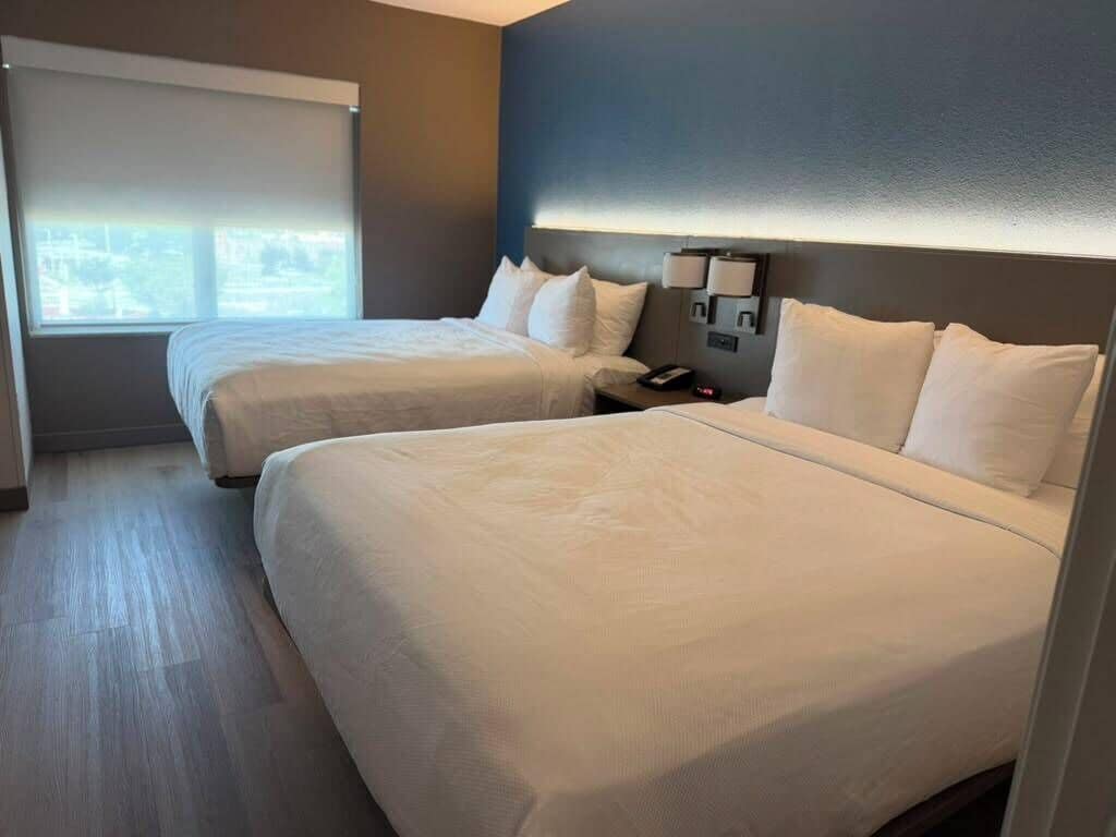 A hotel room with two beds, a nightstand, a lamp, and a window with blinds.