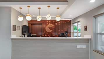A hotel reception desk with a modern design, featuring a wooden panel wall with a crescent moon motif, a flat-screen TV, a cash register, and a digital kiosk.