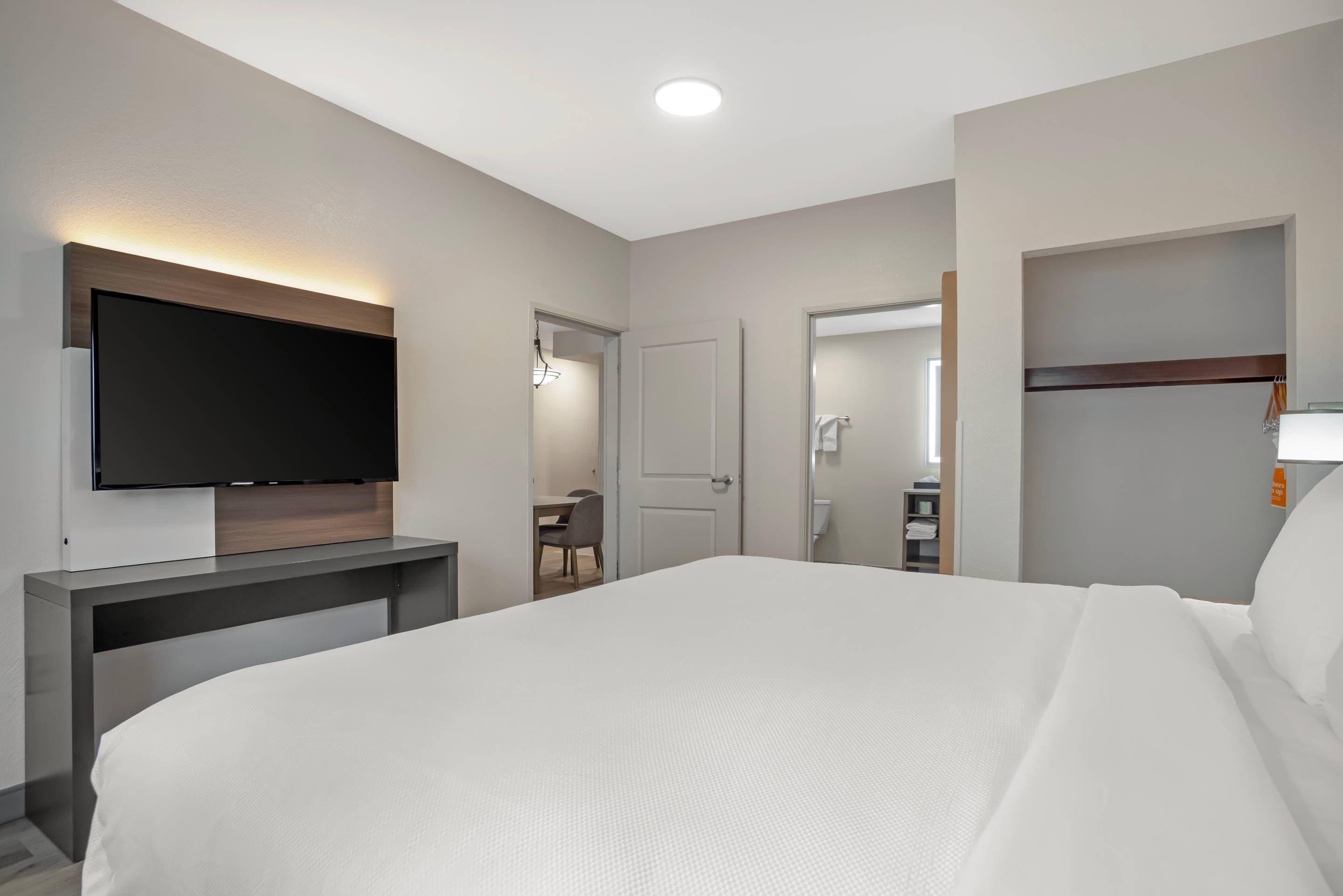 A hotel room with a bed, a flat-screen TV mounted on a wooden console, a desk with a chair, and a bathroom visible through an open door.