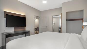 A hotel room with a bed, a flat-screen TV mounted on a wooden console, a desk with a chair, and a bathroom visible through an open door.