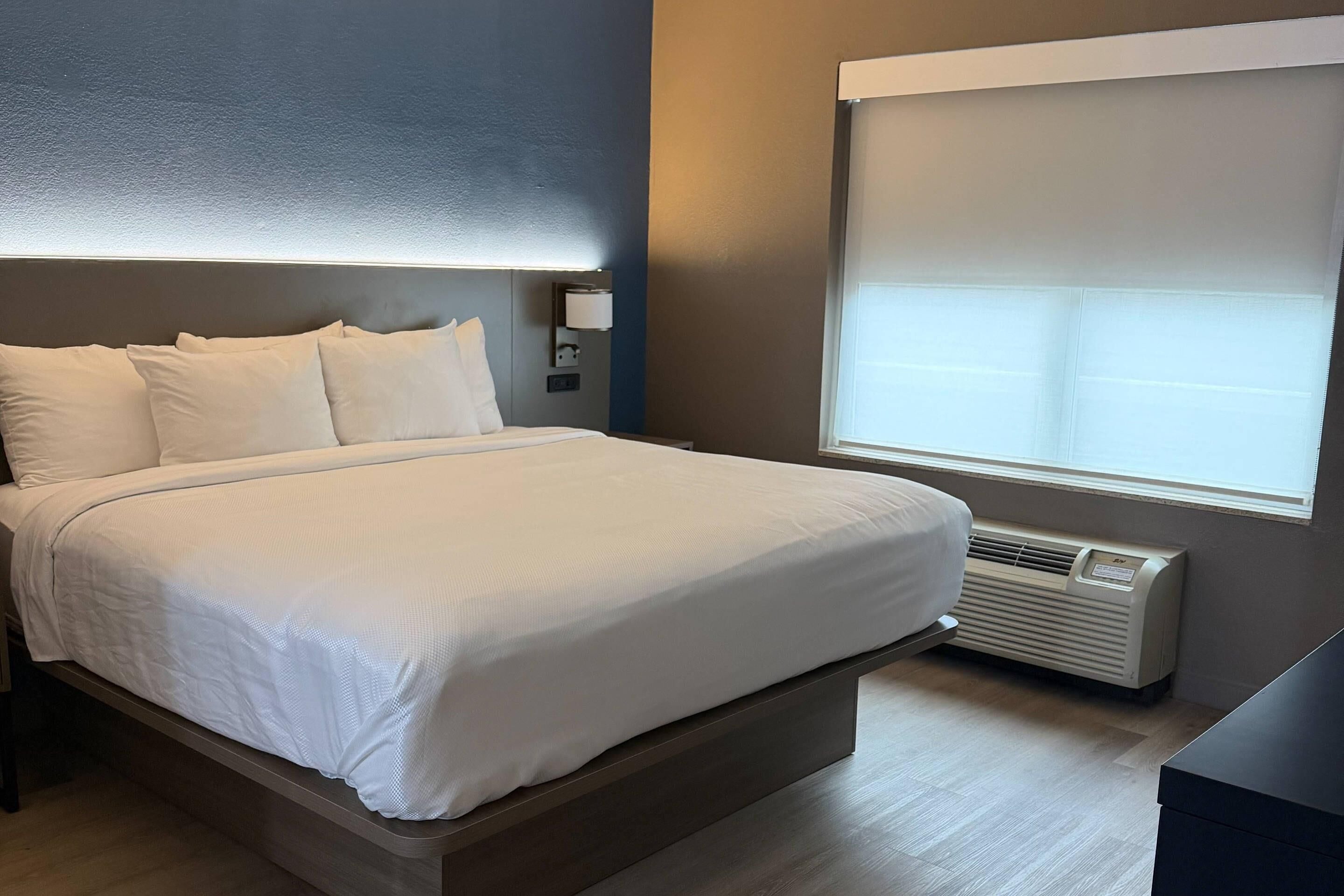 A hotel room with a bed, bedside table, lamp, air conditioning unit, and a window with blinds.