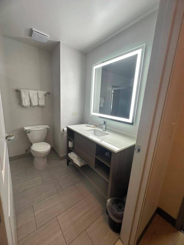A small bathroom with a toilet, a sink, a mirror with built-in lighting, and a trash bin.