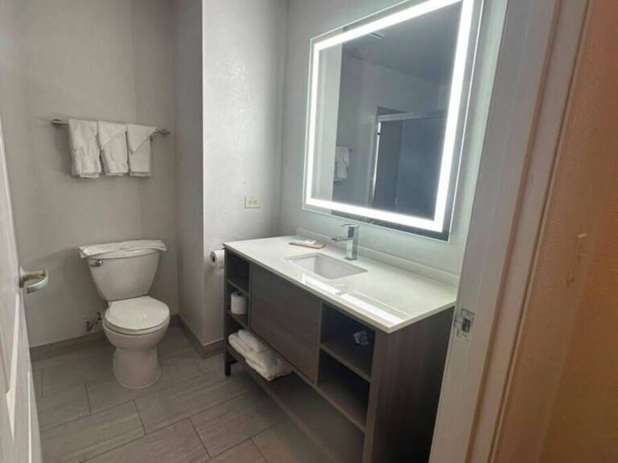 A small bathroom with a toilet, a sink, a mirror with built-in lighting, and a trash bin.