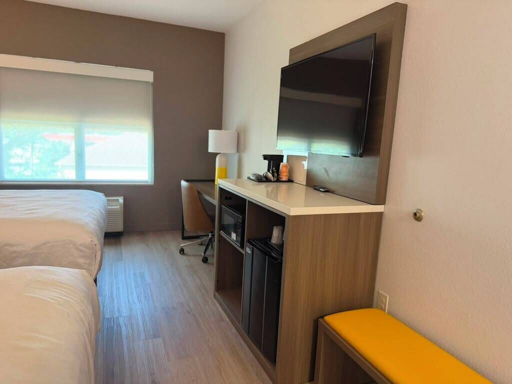 Une chambre d’hôtel avec un lit, un bureau avec un ordinateur, une télévision et une fenêtre avec des stores.