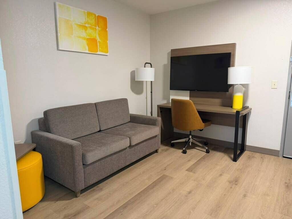 A modern room with a grey sofa, a yellow ottoman, a desk with a lamp, and a TV.