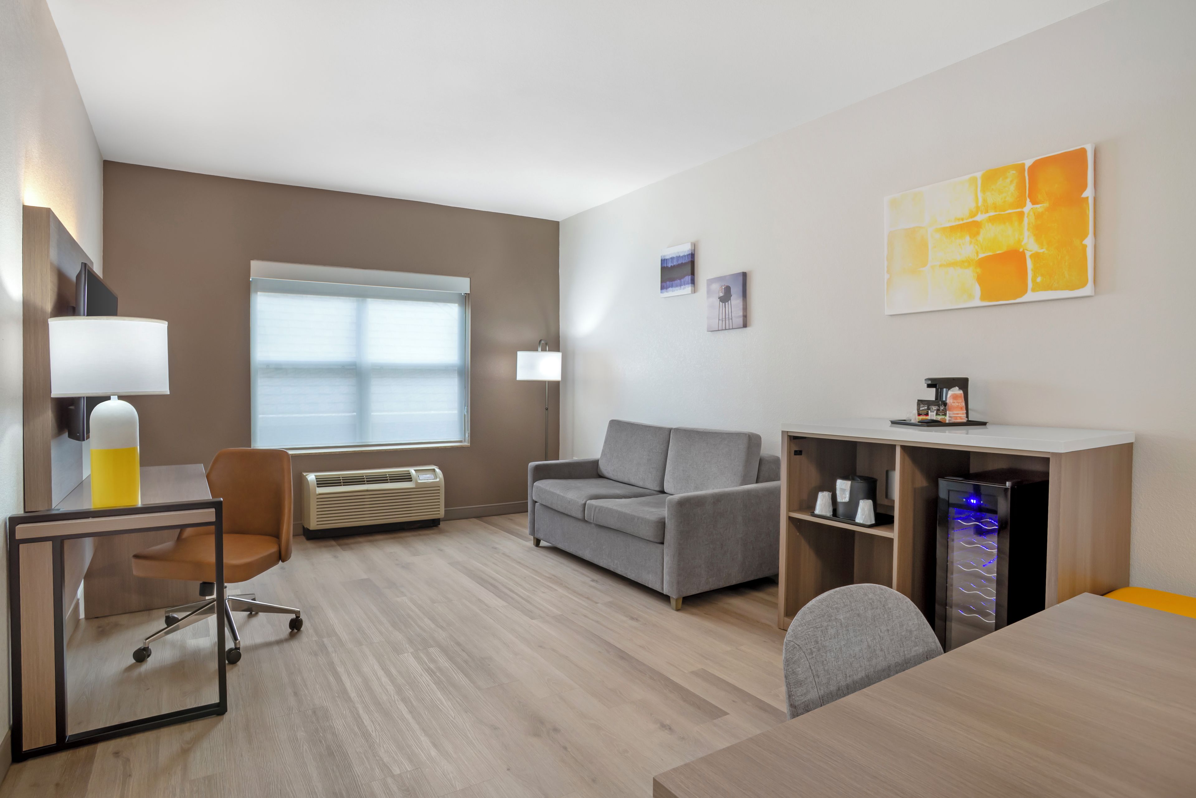 Una habitación de hotel moderna con escritorio, silla, sofá y una pequeña zona de kitchenette.