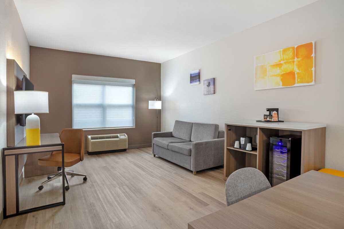 A modern hotel room with a desk, chair, sofa, and a small kitchenette area.