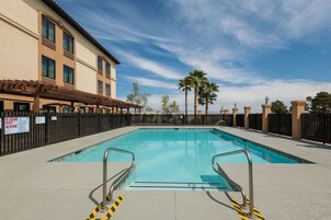Una piscina con barandillas de seguridad, rodeada por una valla, y un edificio con varias ventanas y una pérgola.