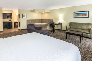 A hotel room with a large bed, a sofa, a chair, a small table, a desk, a lamp, a painting, a bathroom with a bathtub, and a kitchenette.