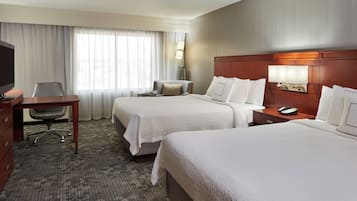 A hotel room with two beds, a desk, a chair, a TV, and a lamp.