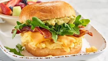 A breakfast sandwich with avocado, bacon, and cheese on a bun, served on a plate with a side of fruit.