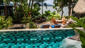 Una piscina con mosaicos, rodeada de sillones de madera y plantas tropicales.