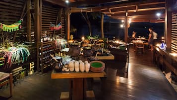 An outdoor dining area with wooden tables and chairs, a variety of dishes and drinks, and a view of the night sky.