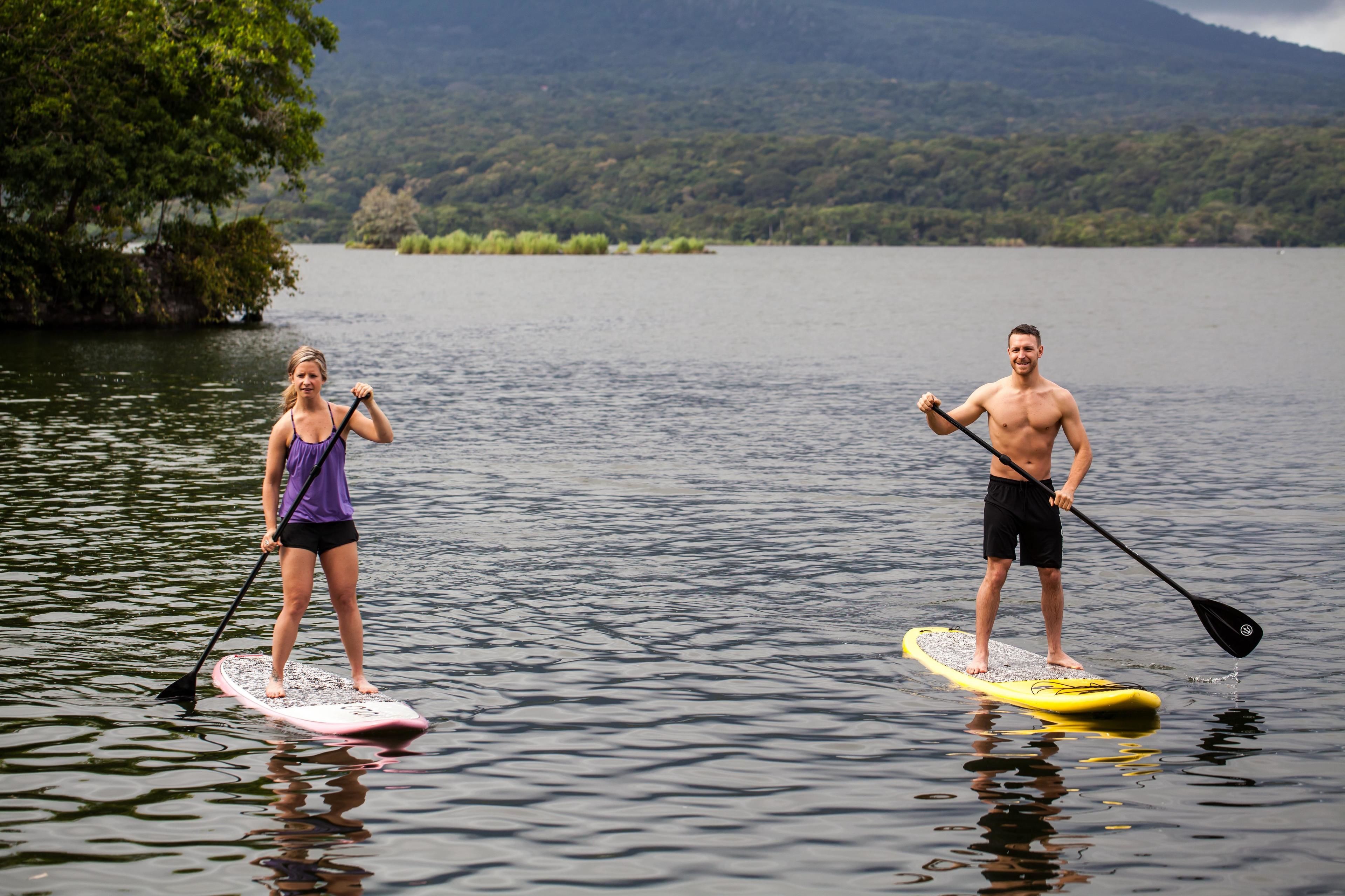 Two people paddleboarding on a calm lake with a forested mountain in the background.