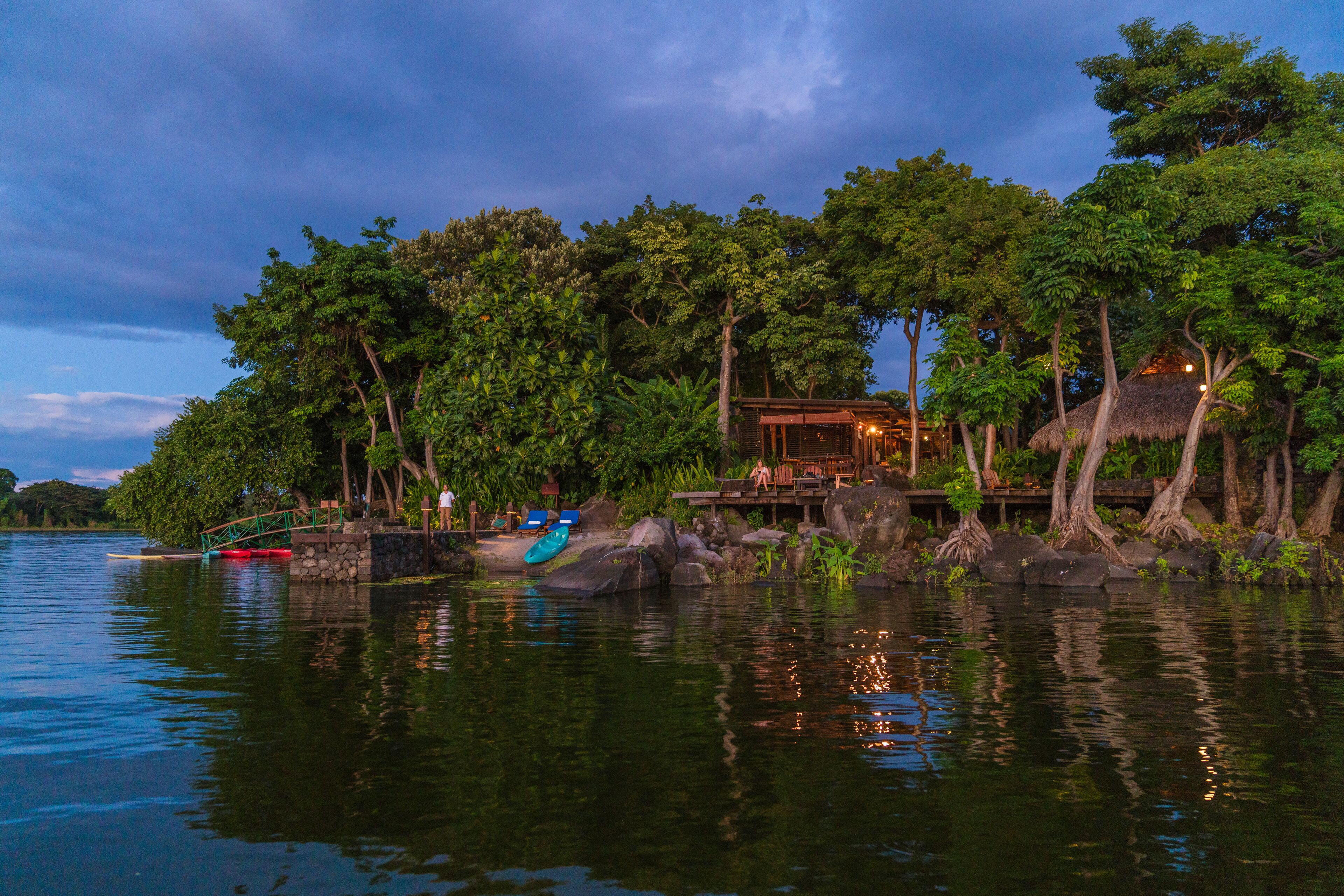 A lakeside resort with a thatched-roof building, kayaks, and a wooden deck overlooking the water.