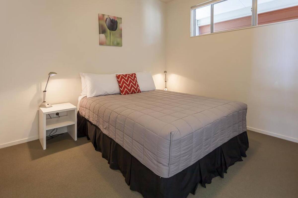 A bedroom with a bed, a nightstand, a lamp, a window, and a wall-mounted picture.