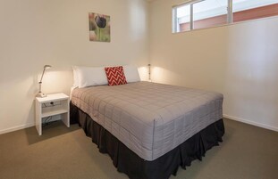 Un dormitorio con cama, mesita de noche, lámpara, ventana y un cuadro colgado en la pared.