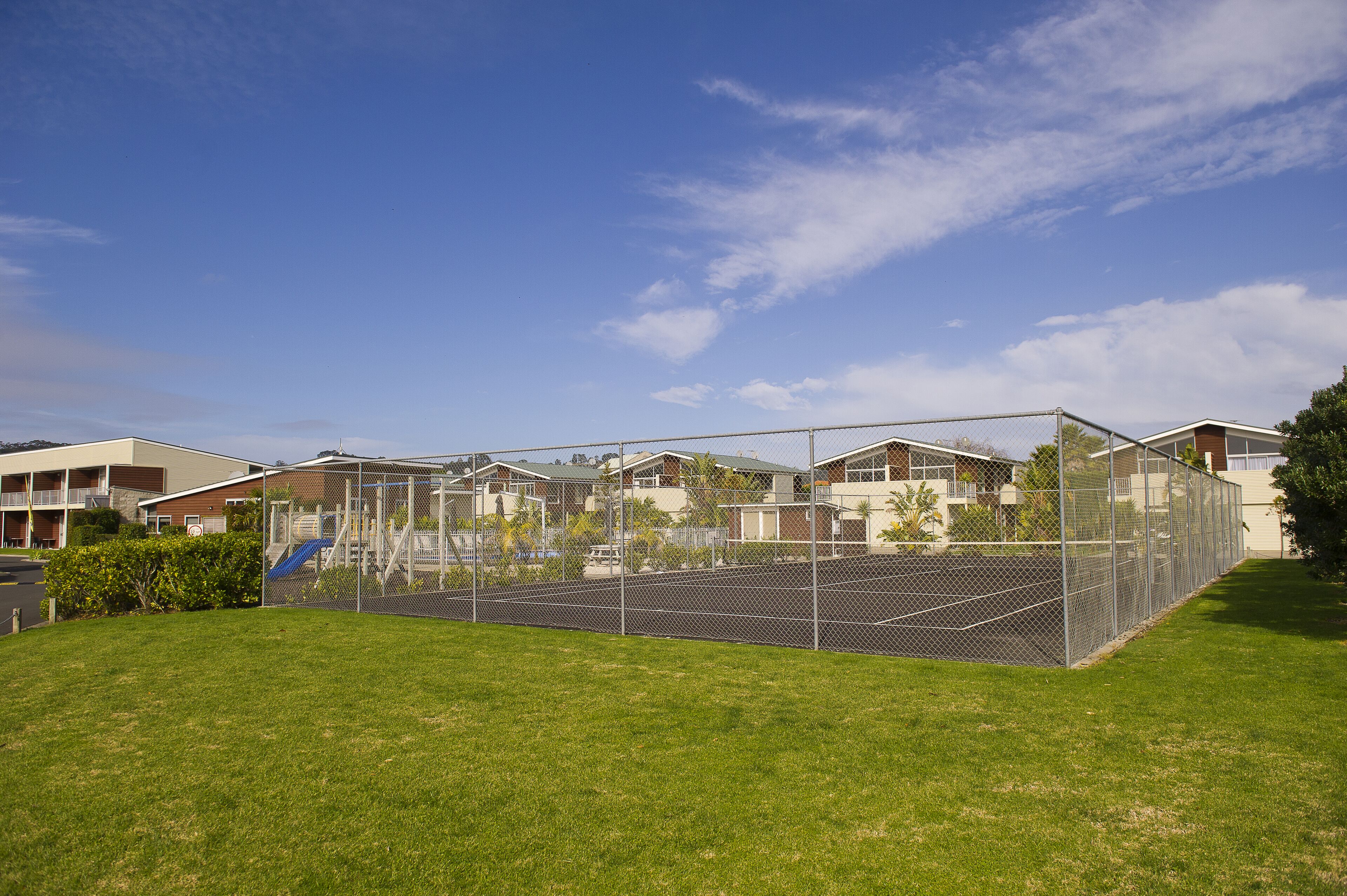 A fenced tennis court with a grassy area in front, surrounded by residential buildings.