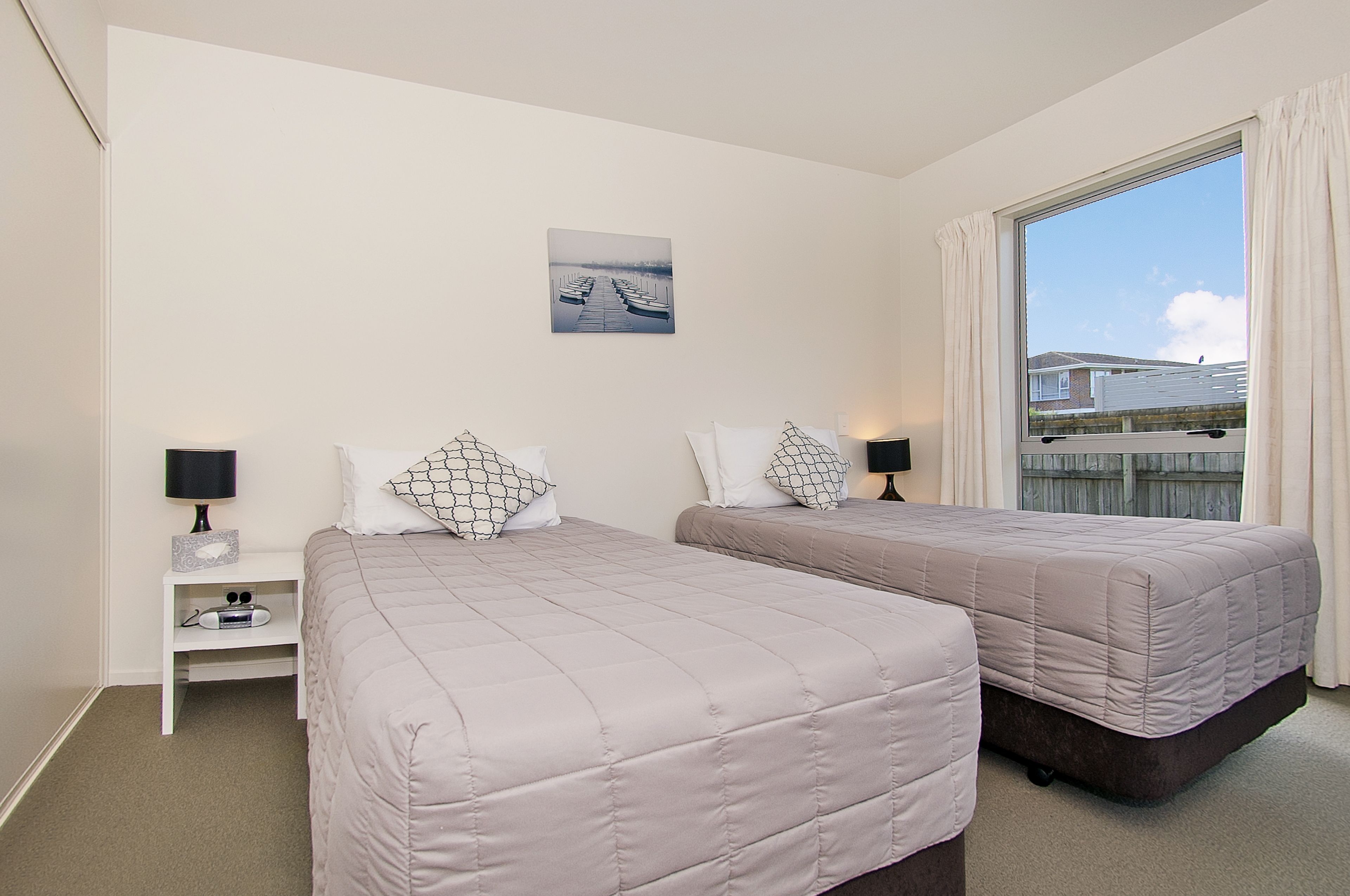 A hotel room with two beds, a nightstand with a lamp, a small shelf, and a window with curtains.