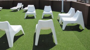 Une terrasse sur le toit, avec des chaises en plastique blanc et du gazon synthétique, offrant une vue sur un paysage urbain composé d’immeubles et d’un ciel dégagé.