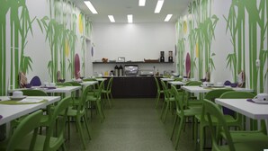 Un espace repas avec des chaises vertes et des tables blanches, un coin cuisine équipé d’appareils électroménagers, et un mur orné de motifs verts et jaunes.