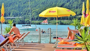 A lakeside pool area with lounge chairs and umbrellas, a boat, and a house in the background.