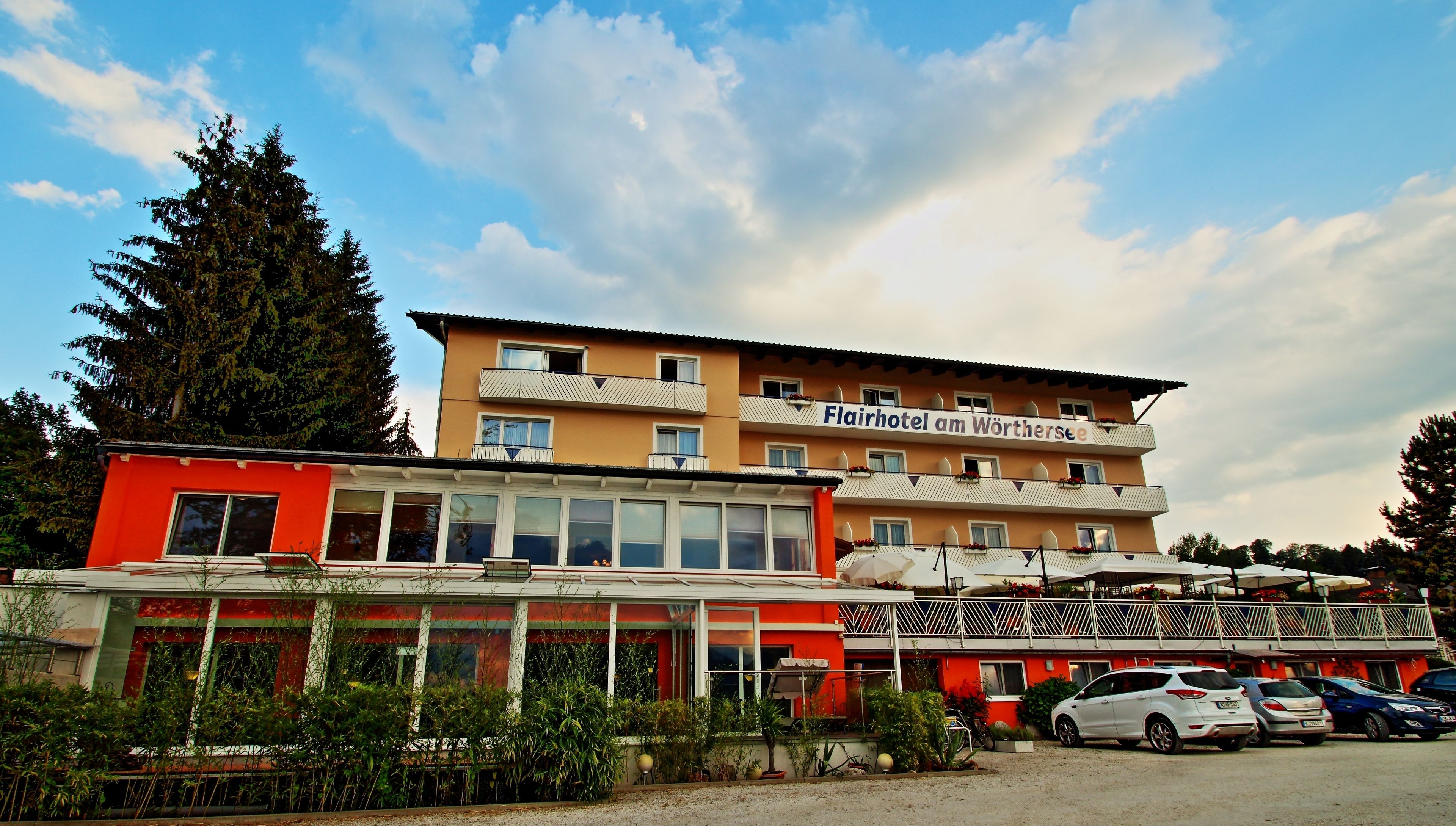 A hotel with multiple balconies, large windows, and a parking area with several cars.