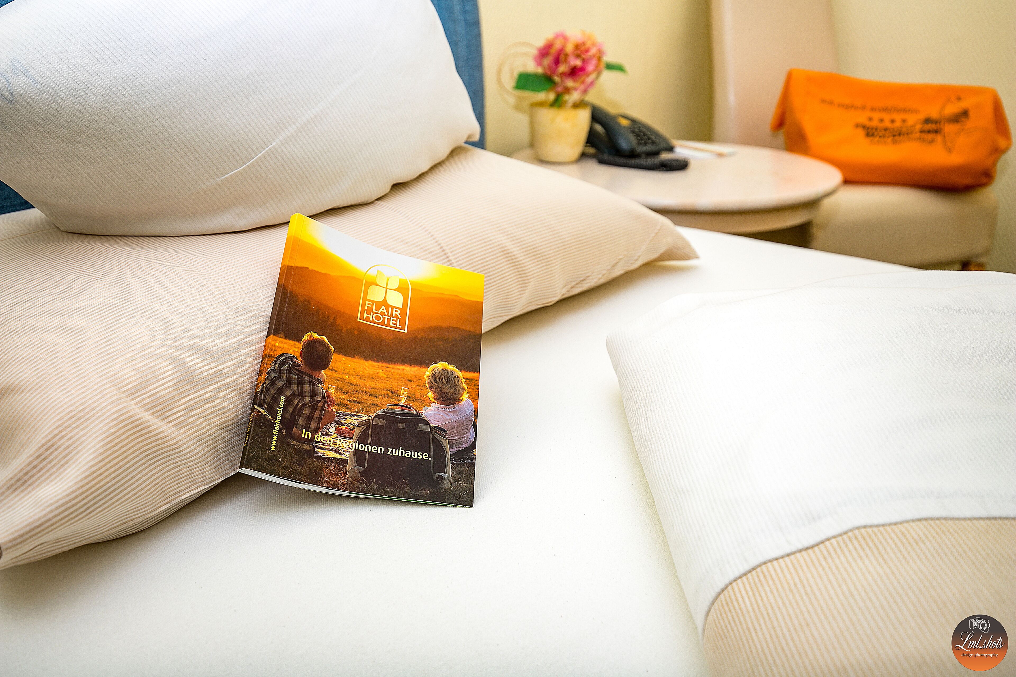 Ein ordentlich gemachtes Bett mit Kissen, eine Zeitschrift auf dem Bett, ein kleiner Tisch mit Telefon und Blumenvase sowie ein Stuhl mit orangefarbenem Kissen.