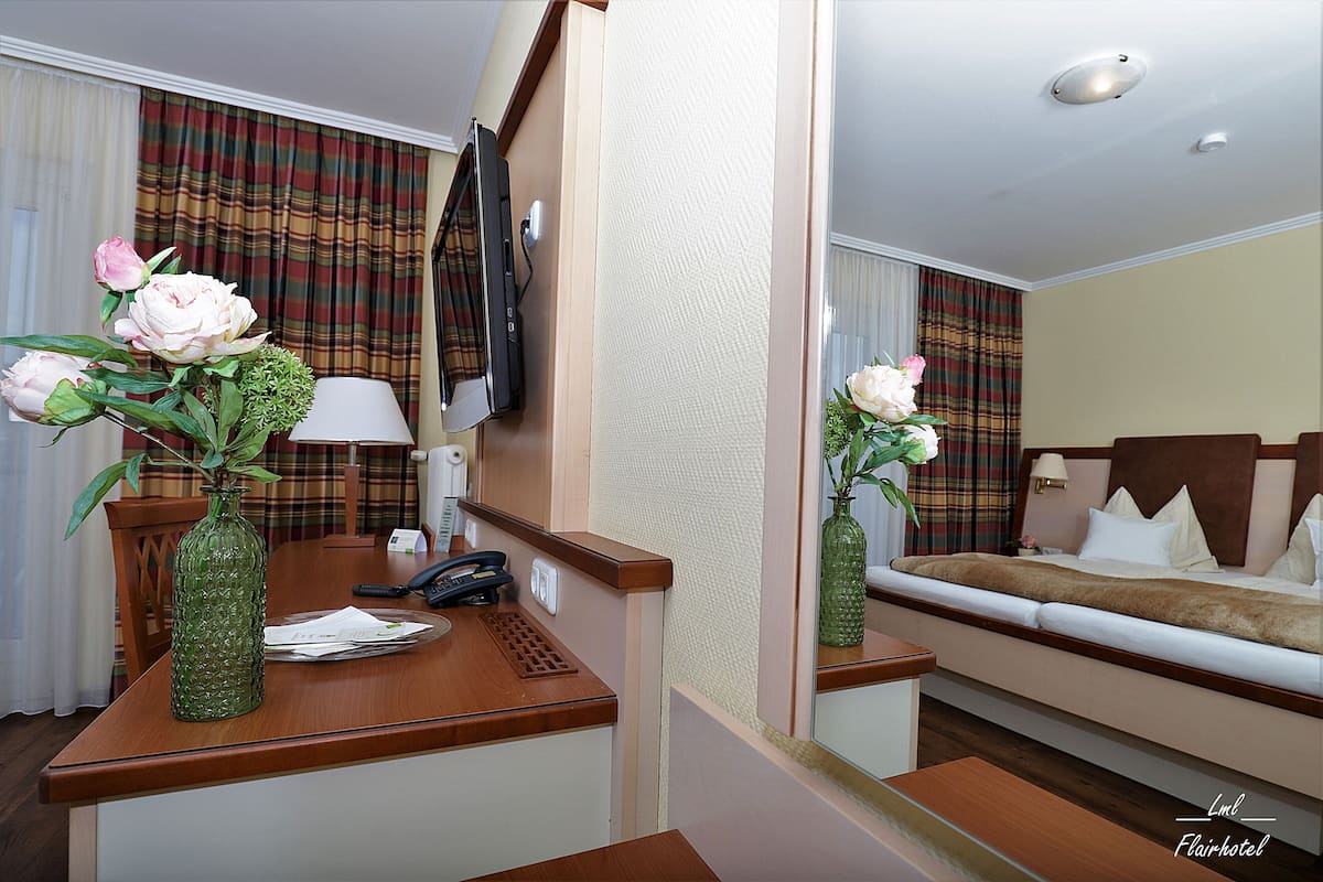 Hotelkamer met een houten bureau, telefoon, spiegel, een bed met wit beddengoed en twee vazen met bloemen.