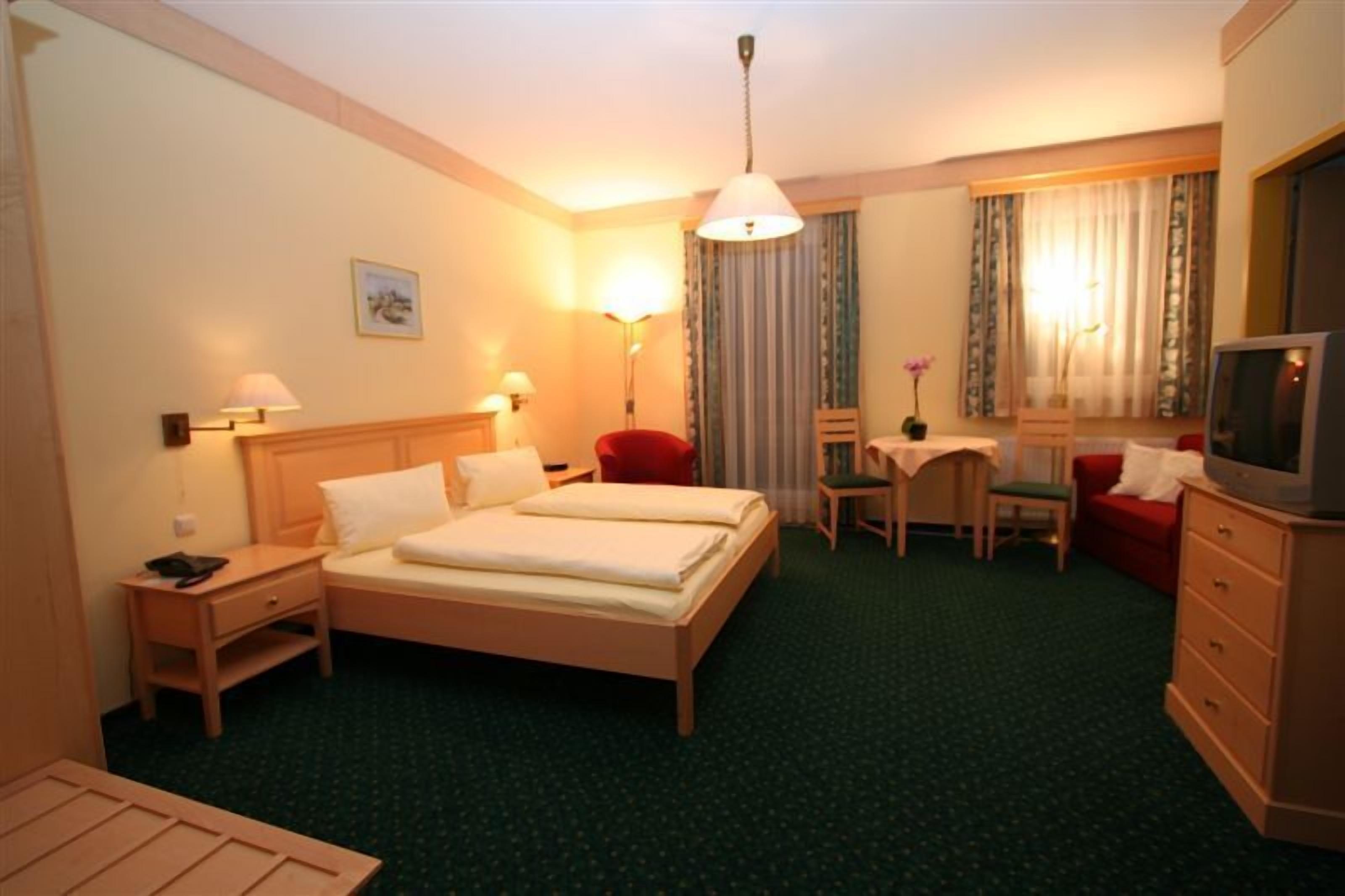 Una habitación de hotel con cama, mesitas de noche, un escritorio con silla, televisión y ventana con cortinas.