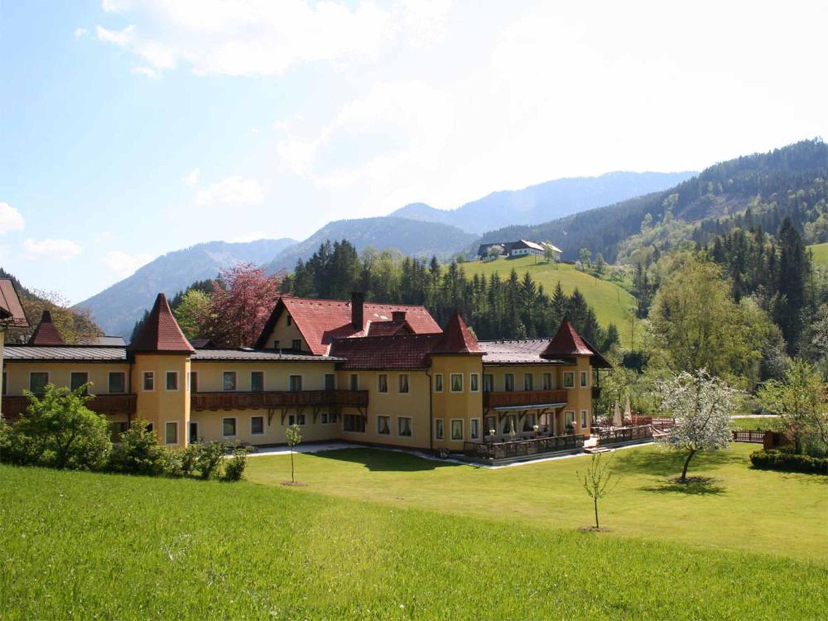 A large yellow building with red roofs, surrounded by green fields and mountains in the background.