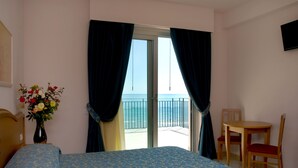 Un cuarto de hotel con cama, una mesita con dos sillas, un jarrón de flores y un balcón con vista al mar.