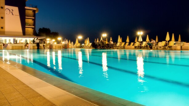 Una piscina con sillas de descanso y sombrillas, rodeada por un edificio con un logotipo visible.