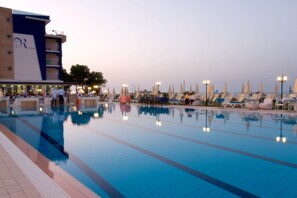 A swimming pool with lounge chairs and umbrellas, a building with a large logo, and a clear sky. - Roses Hotel (Roseto degli Abruzzi)