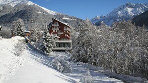 Un edificio de estilo chalet con fachada roja y balcones, rodeado de árboles nevados y montañas al fondo.