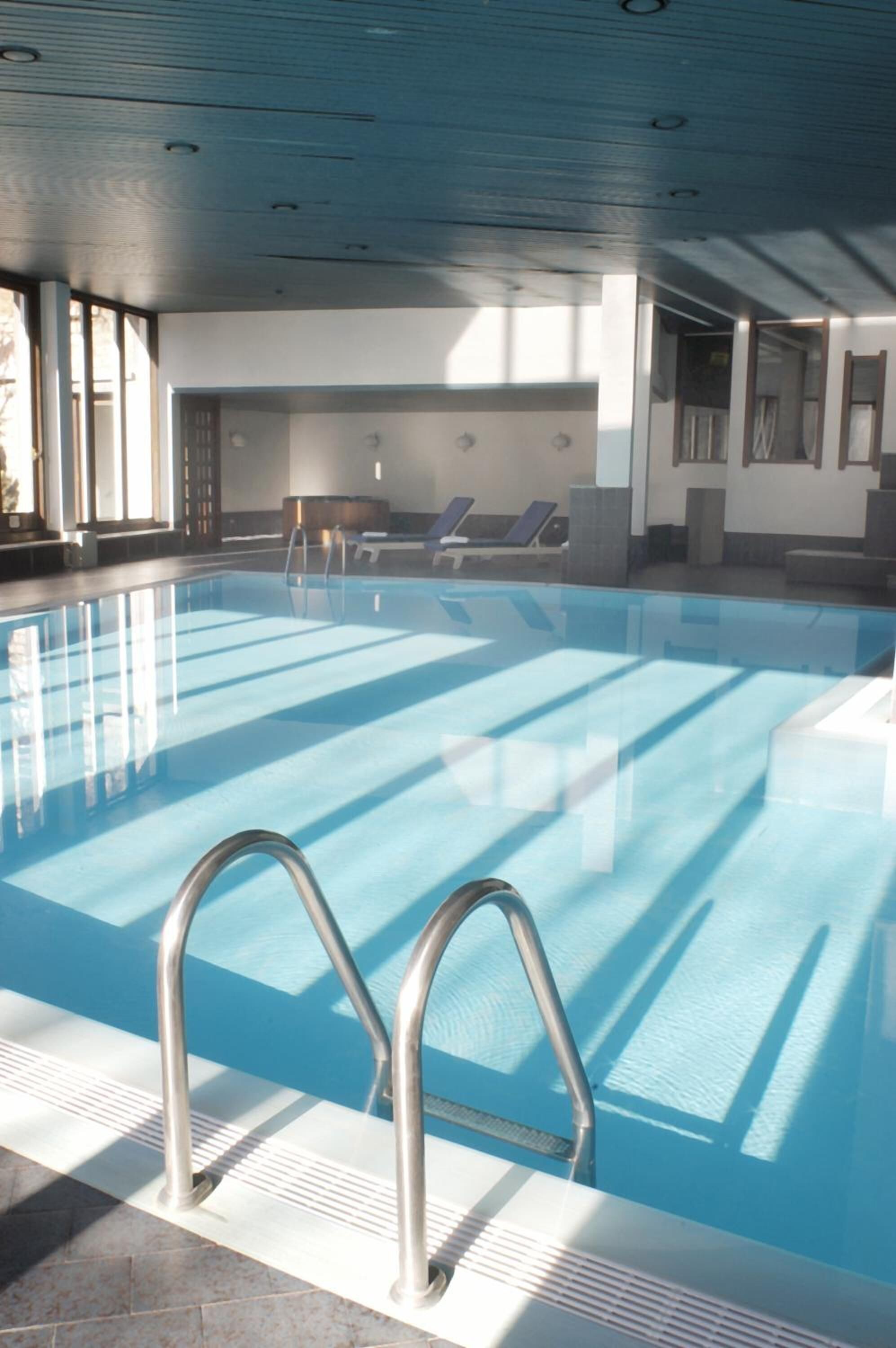 An indoor swimming pool with clear water, a metal ladder, and sunlit lounge chairs.